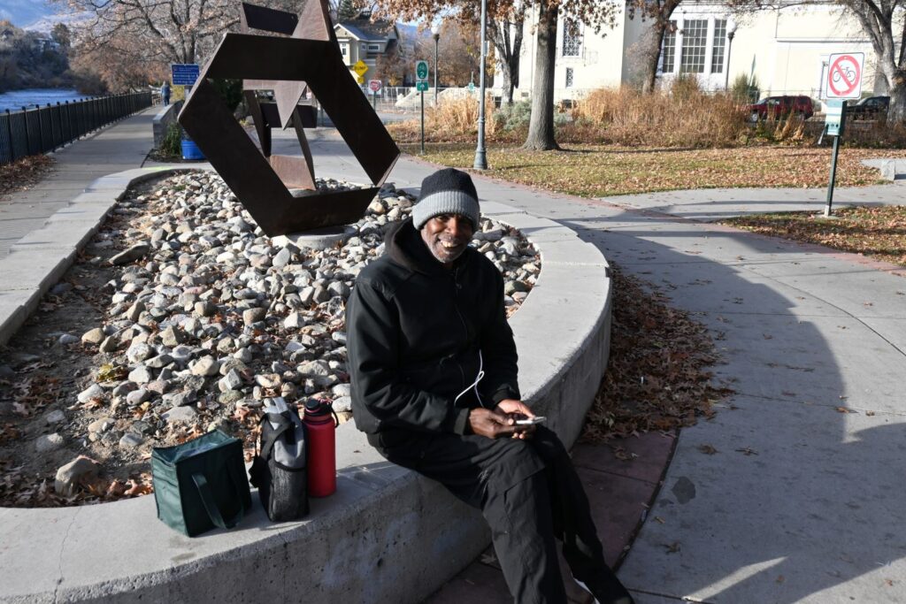 A man in winter clothing sits bundled underneath a sculpture by the Truckee River.