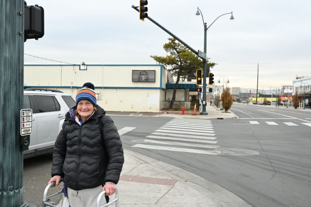 A woman with a walker stands on a street corner smilin.